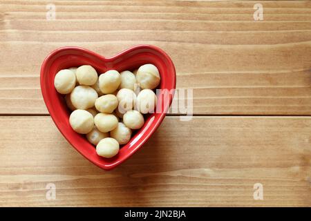 Macadamia nuts lie in a heart-shaped ceramic bowl, which stands on a wooden table. View from above Stock Photo