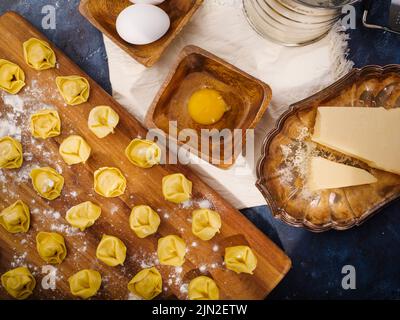 The process of making homemade Italian ravioli. small raw ravioli on a wooden cutting board, ingredients and kitchen utensils on a dark blue backgroun Stock Photo