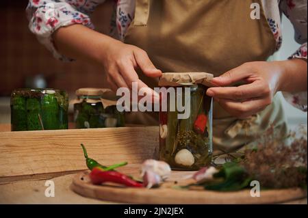 Female hands putting craft paper on the lid covering a glass jar of homemade pickled chili peppers. Pickling. Canning Stock Photo