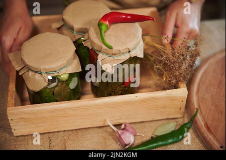Cropped view of a wooden crate with jars with delicious homemade tasty pickled cucumbers and marinated chili peppers Stock Photo