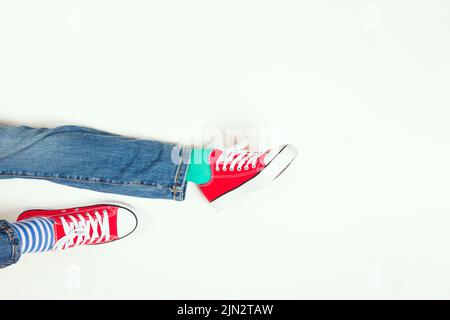 Teenage boy wearing different pair of socks. Child foots in mismatched socks and red sneakers sitting on white background. Odd Socks day, Anti Stock Photo