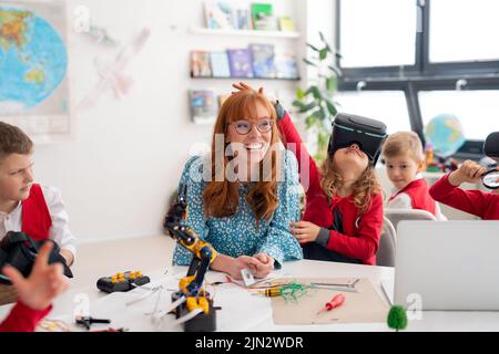 Happy schoolgirl wearing virtual reality goggles at class, having fun with teacher and classmates. Stock Photo
