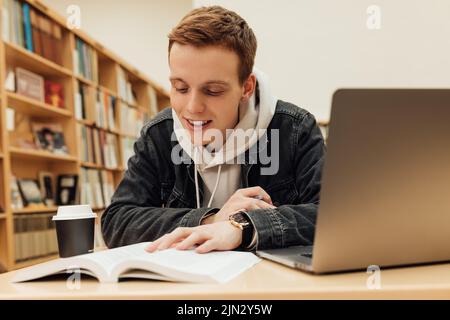Smiling student with ginger hair preparing exams in library. Student reading from book while sitting at desk. Stock Photo