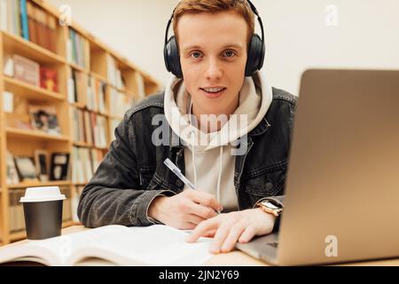 Smiling student in headphones holding pen sitting at desk looking at camera Stock Photo