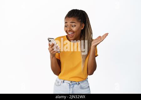 Image of happy black girl reacting surprised at phone notification, looking at smartphone and rejoicing, celebrating and smiling, standing over white Stock Photo
