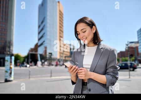 Young smiling Asian business woman using cellphone standing on city street. Stock Photo