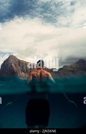 A vertical shot of a back-standing man waist-deep in the water against beautiful mountains under the blue cloudy sky Stock Photo