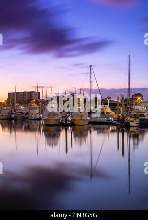 A vertical shot of a harbor in the evening with boats on the water, long exposure shot of clouds, sunset sky and a city in the background. Stock Photo