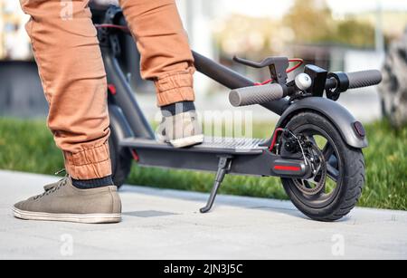 Man feet on parked electric scooter, closeup detail, sun lit blurred grass in background Stock Photo
