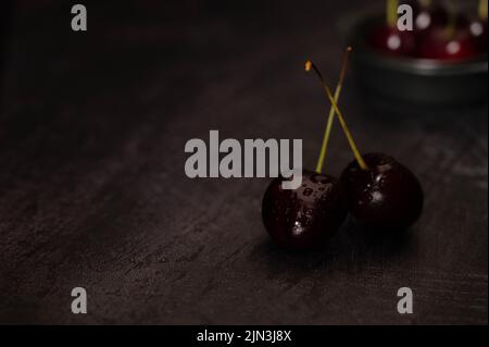 Dark atmospheric image of two ripe red cherries with crossed stems and a ceramic bowl full of cherries in the background on a dark metalic background Stock Photo