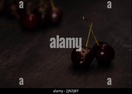 Dark atmospheric image of two ripe red cherries with crossed stems and a pile of cherries in the background on a dark metalic background with copyspac Stock Photo