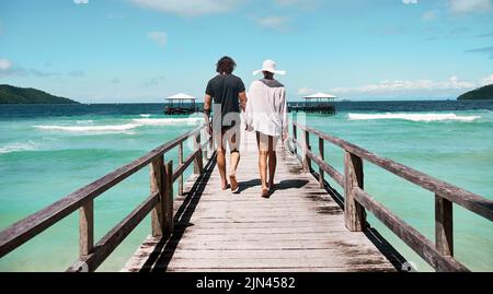 I enjoy our little walks. Rearview shot of a carefree young couple walking on a jetty together next to the ocean during the day. Stock Photo