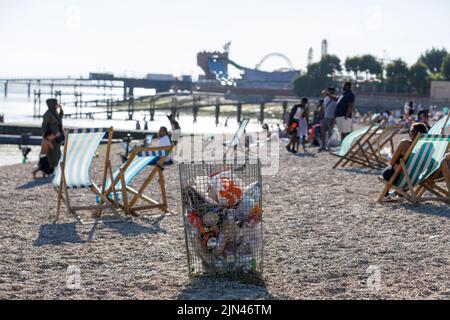 A trash can on a beach with deck chairs and people blurred in the background on a hot sunny day in summer. Rubbish bin. Stock Photo