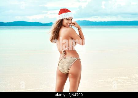 I hope santa knows how good Ive been. a young woman wearing a christmas cap while standing on the beach. Stock Photo