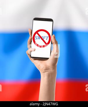 Hand holding smartphone with crossed out emblem of SWIFT on screen against Russian flag. Concept of Ukraine-Russia-related sanctions Stock Photo