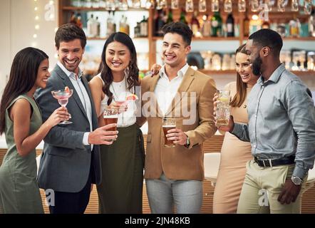 We need to start doing this more often. a group of young friends holding their drinks while standing in a bar. Stock Photo