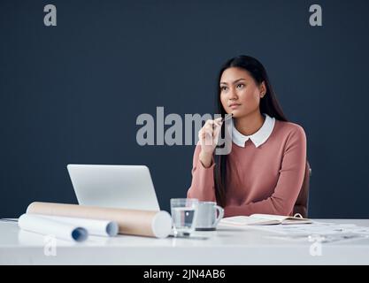 Shes got a mindful of ideas. an attractive young female architect looking thoughtful while working on a laptop in her office. Stock Photo