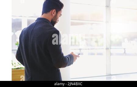 My work never ends. Rearview shot of a handsome young businessman standing and using his cellphone while pulling a suitcase in the office. Stock Photo