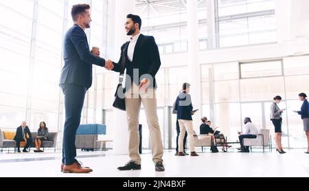 Its so good seeing you again. Full length shot of two handsome young businessmen shaking hands while their colleagues work behind them in the office. Stock Photo