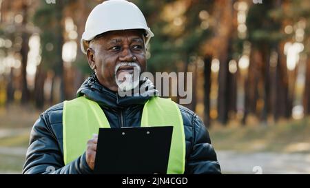 Mature concentrated forestry engineer technician forester in protective helmet stands in park assesses situation makes plan for cutting trees observes Stock Photo
