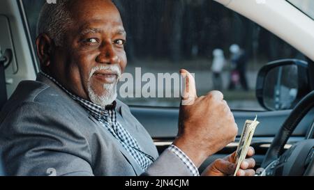 Elderly male sitting in car counting cash payment wages mature african american businessman calculates income successful business happy smiling man Stock Photo