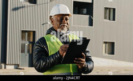 Elderly african american man construction manager evaluates building old foreman standing on street in protective hardhat writes data to tablet Stock Photo