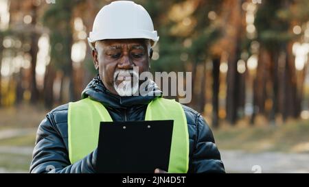 Mature concentrated forestry engineer technician forester in protective helmet stands in park assesses situation makes plan for cutting trees observes Stock Photo