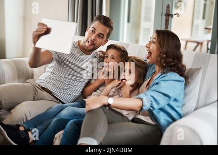 Silly selfies are the best selfies. a young family of four taking selfies together with a digital tablet on the sofa at home. Stock Photo
