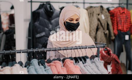 Young arab woman shopper in medical protective mask chooses clothes in clothing store retail sales discount purchase shopping in boutique muslim Stock Photo
