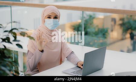 Young arab woman freelancer in medical mask sitting at workplace working typing on laptop checking new computer app software leaving good feedback Stock Photo