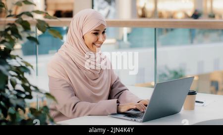 Happy young arab businesswoman sitting at office desk typing on laptop working on internet checking new app smiling looking at camera showing thumb up Stock Photo