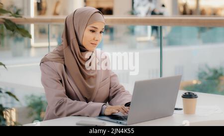 Focused serious arab businesswoman professional entrepreneur sitting at office desk typing looking at laptop screen studying online course muslim girl Stock Photo