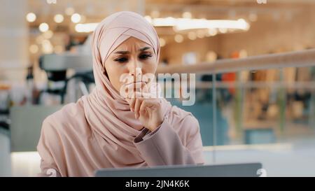 Young pensive puzzled arab woman in hijab working on laptop concentrating on idea looking for inspiration keeps hand on chin thinking deeply in Stock Photo