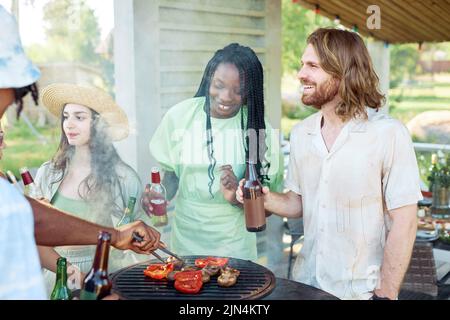 Cheerful group of friends grilling meat and vegetables while enjoying barbeque party outdoors in Summer Stock Photo