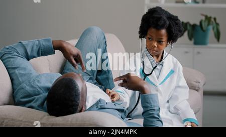 Cute little girl in medical gown plays with dad pretending to be doctor man lying on sofa funny daughter listening to patient father in room holds Stock Photo