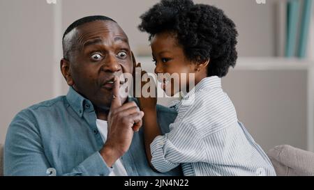 Little african american daughter whispering secret adult dad in ear telling gossip surprised dad carefully listens confidential information making Stock Photo