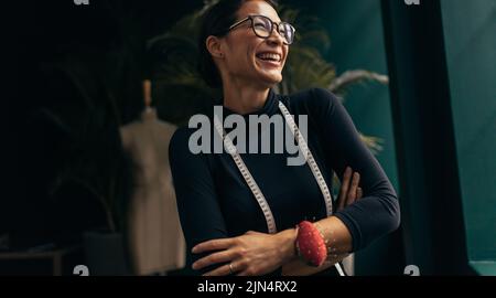 Laughing female fashion designer in her studio. Asian woman standing with measuring tape around neck and looking away. Stock Photo