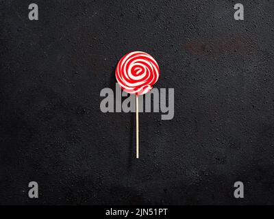 Red and white colored swirl round candy lollipop on black textured background. Stock Photo