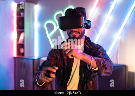 Young man on neon background playing video game virtual reality or VR headset at home using gamepads - concepts of metaverse, cyberspace and Stock Photo