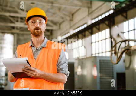 White man wearing helmet and vest writing on clipboard while working at factory Stock Photo