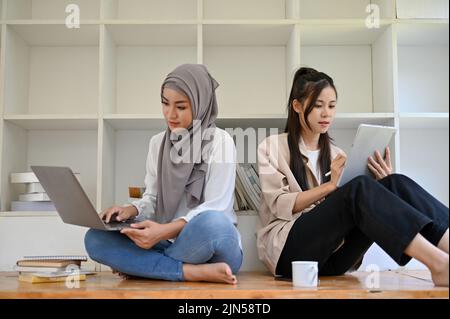 Attractive young Asian Muslim female college student and her Asian female friend focused, using their laptop and tablet to research information on the Stock Photo