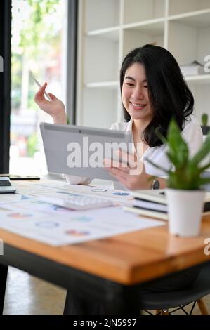 Portrait, Pretty and charming Asian businesswoman or female financial manager using digital tablet touchpad at her office desk. Stock Photo