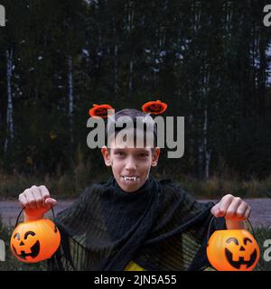 A cheerful boy in vampire costumes shows his teeth holding Jack's lanterns in his hands on a dark background in the park. A child in a Halloween costu Stock Photo