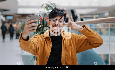 Young handsome arab guy blogger model holding phone in hand photographed on mobile camera posing smiling making gesture taking photo filming video for Stock Photo