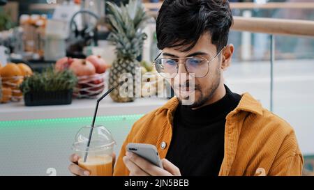 Young happy guy sits in cafe drinks freshly squeezed juice satisfied man holding mobile phone typing message email communicate with friends remote in Stock Photo