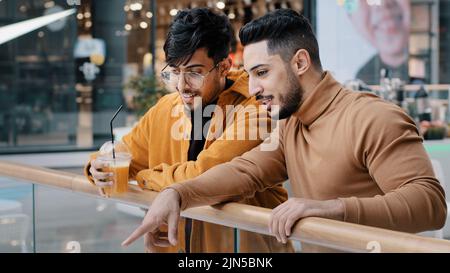 Two positive arab guys friends students stand in public place in shopping and entertainment center talking communicate pointing fingers choose where Stock Photo