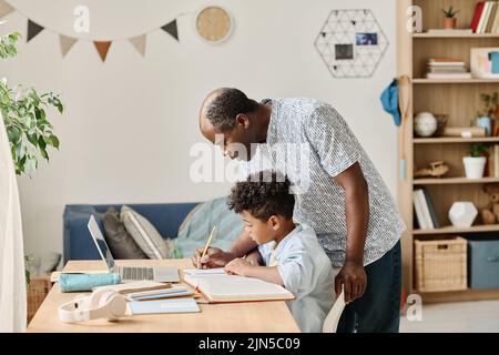 African mature tutor helping schoolboy to do his homework while he sitting at table with books and laptop at home Stock Photo
