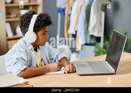 African boy in wireless headphone looking at monitor of laptop and making notes in his notebook during online studying Stock Photo