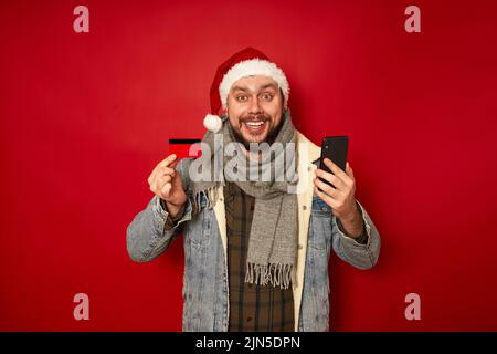 excited man in Christmas hat holds bank credit card and buys phone through mobile application Stock Photo
