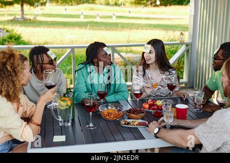 Diverse group of friends playing Guess who game while sitting at table outdoors in Summer Stock Photo